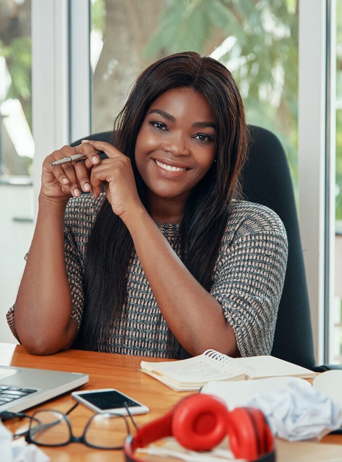 Happy adult black woman sitting at working desk in office smiling at camera adorably