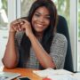 Happy adult black woman sitting at working desk in office smiling at camera adorably